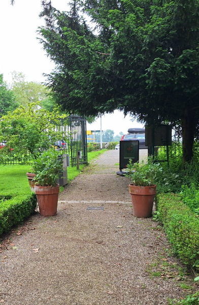 Seen from the garden, a green fence with an open gate. Beyond the fence, cars drive by. On this side you see pots, low hedges, and a tidy gravel path.