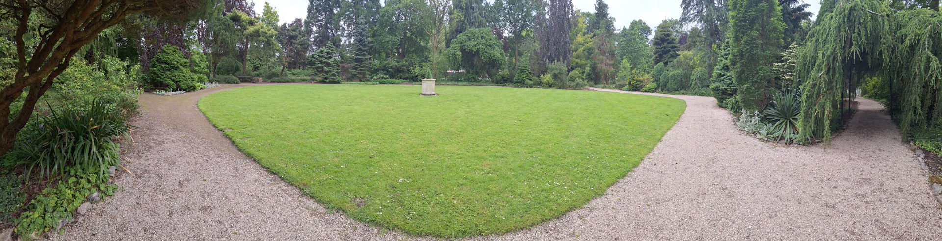 Een groen veld omringd door een cirkelvormig pad. In het midden staat een stenen sokkel zonder beeld. Geen van de paden naar het plein maken een lijn naar het midden.
